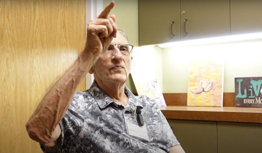 An older man wearing glasses and a patterned shirt gestures with his hand raised, pointing upward, as if emphasizing a thought or memory.